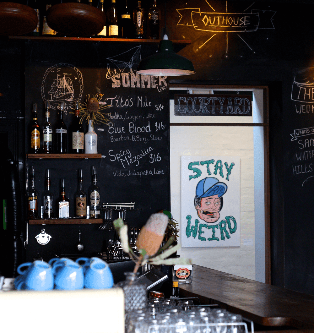 Interior image of coffee machine with cups stacked ontop and some booze bottles on some shelves. In the background a poster with 'stay weird' written.