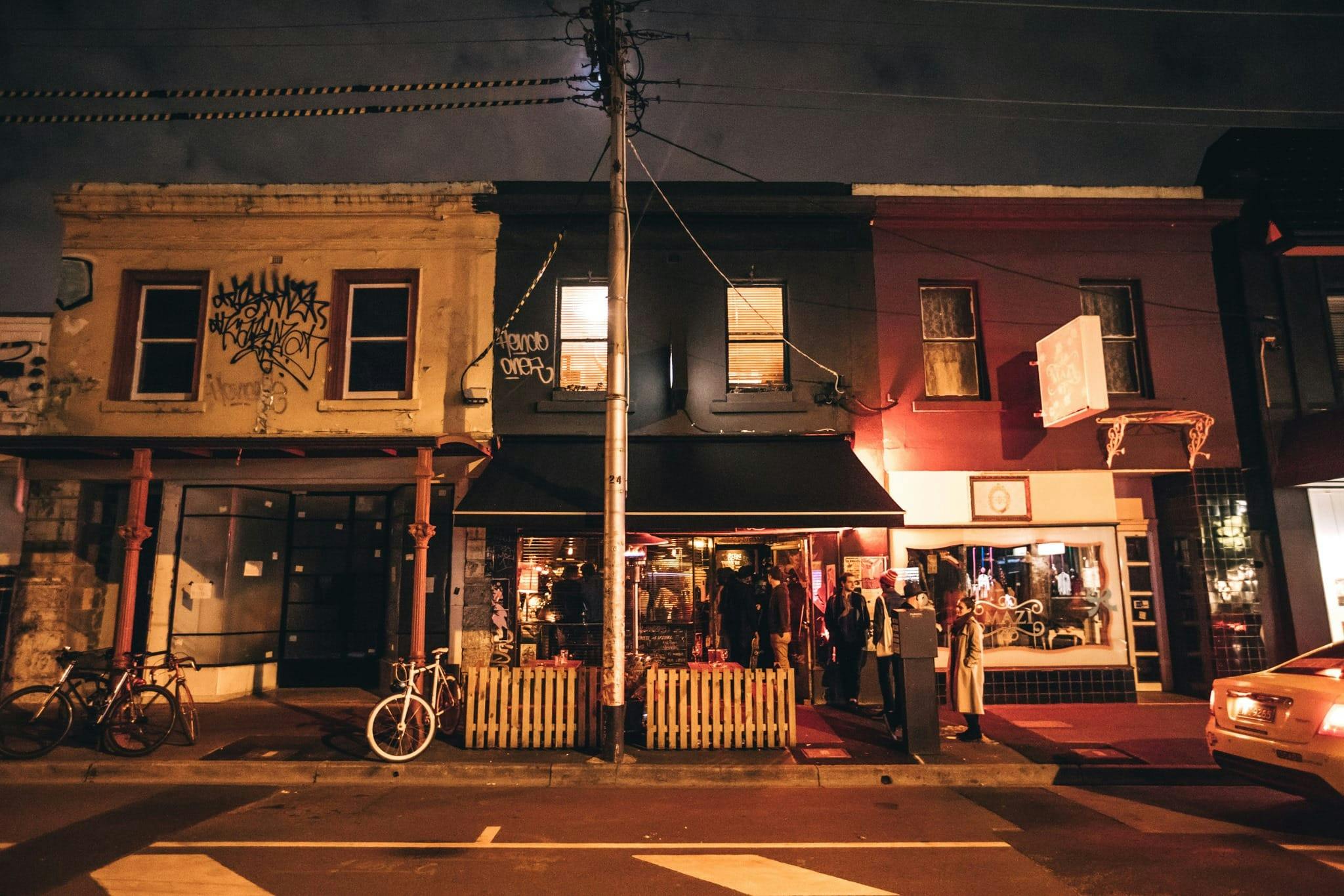Nightime street view of The Rooks Return Bar on Brunswick St, in Fitzroy with groups of people chatting outside, a bike locked up to a power pole and buiolding lit up.