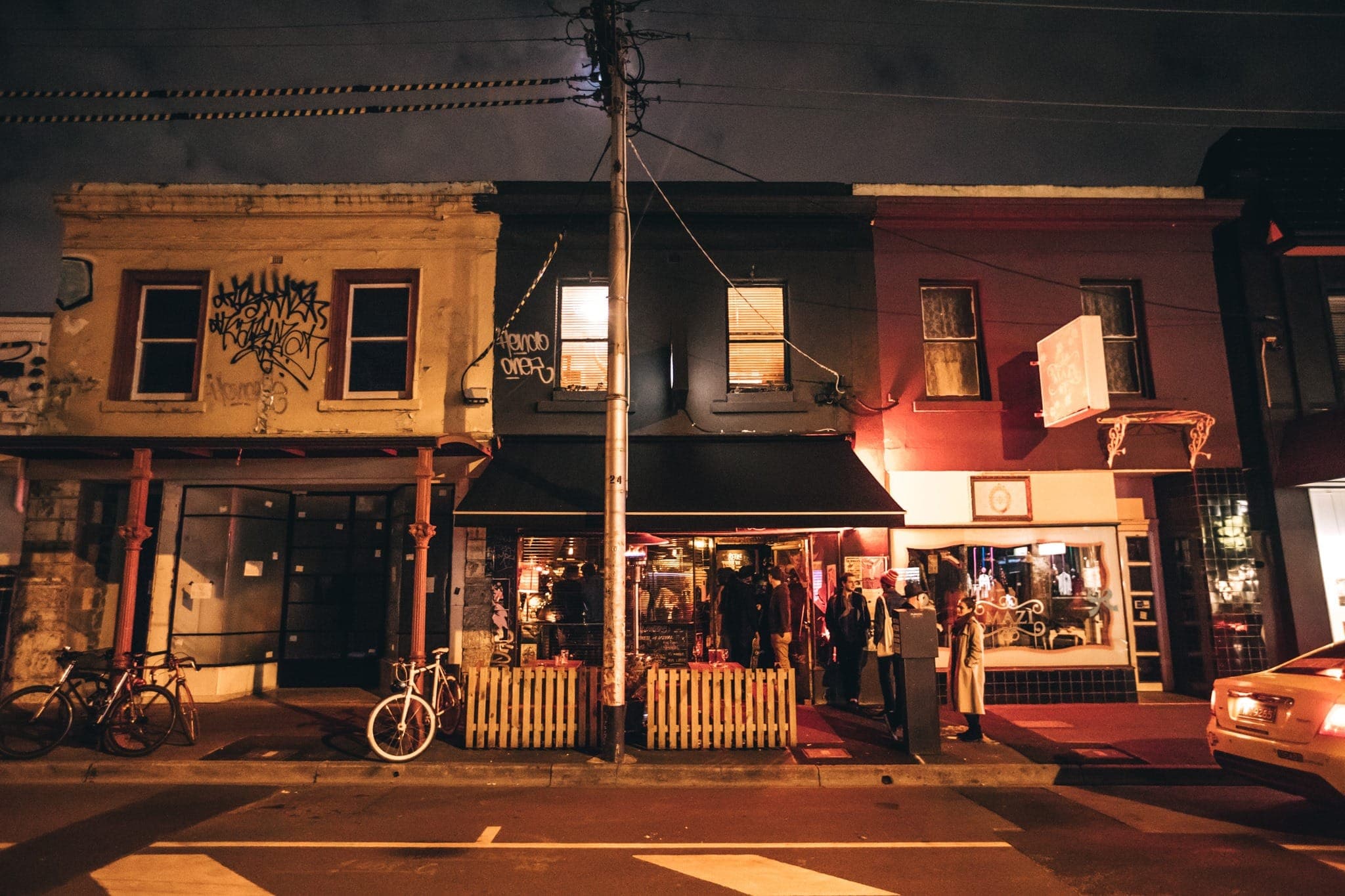 Nightime street view of The Rooks Return Bar on Brunswick St, in Fitzroy with groups of people chatting outside, a bike locked up to a power pole and buiolding lit up.